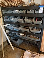 Metal wire shelving unit with gray plastic bins holding assorted hardware items such as plumbing fittings on four shelves. Bottom shelf is empty metal wire.