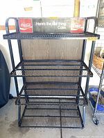 Front view of black metal wire display rack with Coca-Cola branding and four shelves.