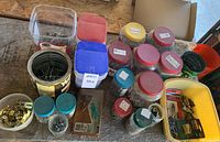 Wide view of multiple plastic and metal containers filled with assorted nuts, bolts, screws, hooks, nails, and metal couplers on a wooden surface.
