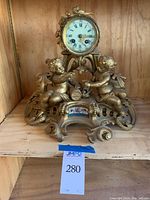 Gold painted ornate mantle clock with cherubs and floral details on wooden shelf.
