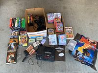 Photo showing Sega Genesis console, two controllers, game cartridges, folded posters, instruction manual, cables, VHS and DVD movies arranged on floor.