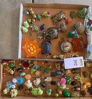 Full view of two cardboard trays containing about 50 tiny turtle figurines in various materials and colors, some with removable lids