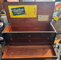Open chest showing cedar lined interior and drawers inside the main compartment, yellow and white label inside the lid showing 'Heirloom Cedar Chest' branding and value features, also certificate sticker inside lid.