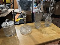 Four crystal items on wooden surface including ice bucket with lid, round stopper decanter, and two wine decanters with elongated stoppers.