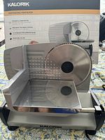 Front and side view of Kalorik food slicer set up with blade, metal carriage, and visible blade guard. Unit is positioned with original box showing product features.