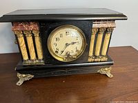 Front view of the mantle clock showing the clock face framed by marble-like columns, black casing, and gold-tone metal feet.
