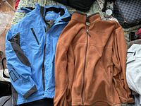 Two men's jackets on a bed, one blue Columbia shell jacket with black accents, one orange Columbia fleece jacket