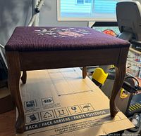 Side view of a needlepoint stool showing wooden legs and floral upholstery on burgundy fabric.