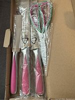 Close-up of the four cutting tools with pink handles still in plastic packaging, showing scissors and three knives.