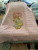 Full view of the pink baby quilt laid out showing the complete applique of the kitten wearing a bonnet, holding a flower and heart, with ruffled edges.
