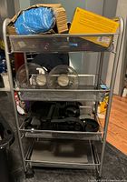 Front and side view of stainless steel rolling cart showing three mesh baskets containing various items, wheels visible with locks on two.