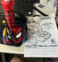 Photo of a red Spider-Man joystick controller with cables and the Spider-Man game instruction manual laid out beside it on a table.
