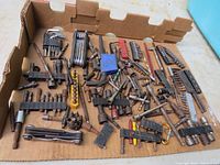 Overview of assorted tool bits and accessories laid out in a cardboard box showing various bits in plastic holders, hex keys, wrench sets, socket adapters, and loose bits; many showing rust.