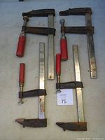 Four large metal bar clamps laid on a flat surface, two pairs with red handles. Clamps show some surface rust and signs of use.