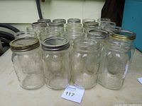 Photo showing 24 clean glass canning jars of 1-liter size including various brands with metal lids on some jars, arranged on an outdoor surface.