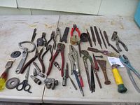 Photo of 47 assorted used hand tools laid out on a table, including pliers, wrenches, files, cutters, scissors, measuring tape, and a mallet. Tools show surface rust and signs of use but appear mostly functional.