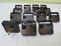 Front angled view of fourteen black plastic quartz clock movement mechanisms arranged in three rows. One mechanism shows brass shaft for clock hands.