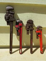 Four used pipe wrenches standing against a yellow exterior wall. Wrenches vary in length and condition showing rust and used wear.