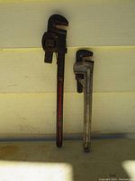 Two pipe wrenches side by side on a pale yellow wall background, one 24-inch steel wrench with red handle and one 18-inch aluminum wrench.