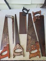 Top-down view showing six different hand saws laid out on a flat surface with mixed handle types and rust on blades