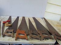 Eight handsaws laid out on a white table, one with a red metal handle and seven with wooden handles. Saw blades show surface rust.