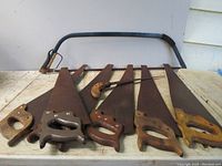Eight handsaws laid out showing various sizes with one large metal handled saw and seven wooden handled saws in varying lengths.
