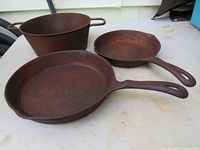 Three vintage cast iron cookware items on a white surface: large skillet with handle, smaller skillet with handle, and a cooking pot with two handles. All show rust and wear.