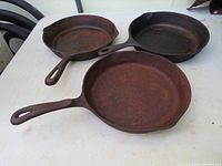 Three rusty cast iron skillet frying pans arranged on a white surface showing general condition and size comparison.