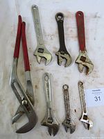Photo showing seven wrenches laid on a white cloth, including two large pipe wrenches with red handles and five adjustable wrenches of various sizes, some with surface rust.