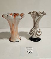 Photo showing two art glass swirl designed vases with ruffled edges, one in white and orange swirls, the other in white and brown swirls, both on black round bases.
