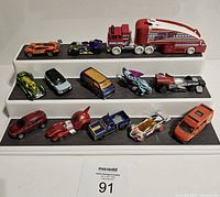 Diecast cars displayed on three black display shelves against a white background. Shows a variety of vehicle types including a large tanker truck, sports and racing cars in multiple colors and styles.