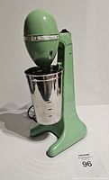 Front-side view of mint green Hamilton Beach drink mixer with stainless steel mixing cup attached. Mixer is plugged in with black cord visible.