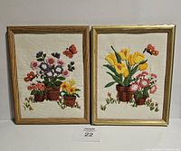 Two framed crewel needlework floral artworks displayed side by side, showing detailed embroidery of flowers in pots and butterflies on a cream colored fabric.