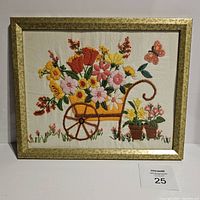 Front view of vintage crewel embroidery showcasing a flower cart filled with colorful flowers and a butterfly, framed in a gold-tone frame.