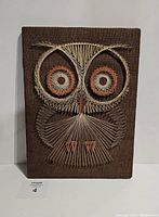 Front view of vintage owl string art on a rectangular board with brown textured fabric background, showing the owl design made of white, orange and brown strings.