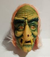 Front view of vintage Topstone rubber ghoul mask showing greenish-yellow textured face, large nose with wart, thick black eyebrows, large yellow fangs, and orange hair around edges.