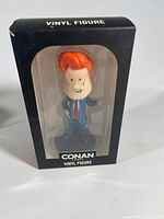 Front view of Conan O'Brien vinyl figure in black packaging showing figure inside, box labeled 'VINYL FIGURE' and 'CONAN VINYL FIGURE'.