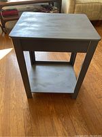 Front side view of gray painted end table showing the flat top surface, drawer front, legs, and bottom shelf on hardwood floor.