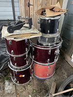 Stack of various drum shells including bass drums and toms with a bass drum pedal on top of a maroon shell and other hardware in cardboard box on black shell.