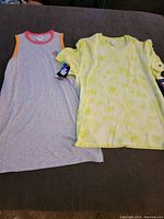 Two new Champion shirts with tags, one sleeveless grey tank top with coral neckline and orange sleeve edges, and one short-sleeve light yellow shirt with puffed sleeves and tie dye pattern.