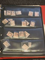 Page of binder showing multiple small old stamps, mostly faded red-orange color, in plastic sleeves on a sheet