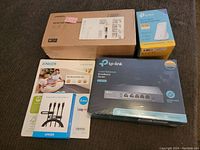 Five sealed electronic items on carpet: UGREEN desktop tablet stand in large box, TP-Link broadband router, TP-Link mesh Wi-Fi system, Anker USB-C adapters and cables.