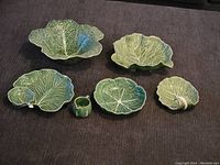 Five green ceramic dishes shaped like cabbage leaves spread out on a surface. Includes two larger leaf bowls, two medium leaf plates, and a small ring-shaped piece. Visible rim chips on two largest dishes.