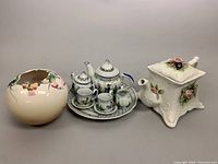 Photo showing a rounded floral porcelain vase with pink and green flowers, a miniature porcelain tea set on a scalloped serving plate, and an embossed floral teapot with a square base and floral decoration.