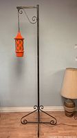 Full view of the tall wrought iron floor lamp with the hanging orange ceramic lamp shade suspended from the bridge arm.