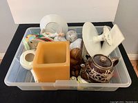 Photo showing miscellaneous chinaware items including plates, teapots, lamps, and decorative pieces packed in clear plastic bin.