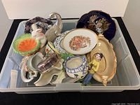 Photo showing assorted bone china plates and figurines in a plastic storage bin including elephant figurine, rabbit, decorative plates, and various ceramic items.