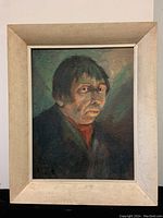 Framed oil painting of a man in a dark coat with reddish shirt, set against a greenish-blue background. The frame is light colored with minor wear.