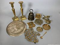Full lot showing all brass items on gray surface including candleholders, bell, plate, duck sculpture, letter B, trivet, dishes, and spoon.