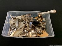 Plastic bin with assorted vintage cutlery and serving utensils including spoons, forks, knives, ladles, and a cake cutter placed inside.